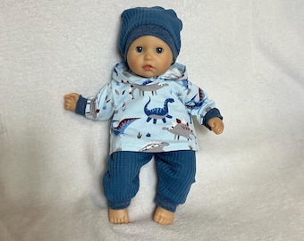 May include: A doll wearing a light blue hooded sweatshirt with a multi-colored dinosaur print, dark blue waffle-knit pants with matching cuffs, and a dark blue waffle-knit beanie. The doll has blue eyes and a neutral expression.