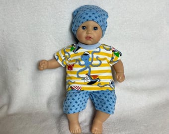 May include: A doll wearing a nautical-themed outfit. The outfit includes a yellow and white striped t-shirt with an octopus design, blue shorts with star patterns, and a matching blue beanie. The doll is on a white background.