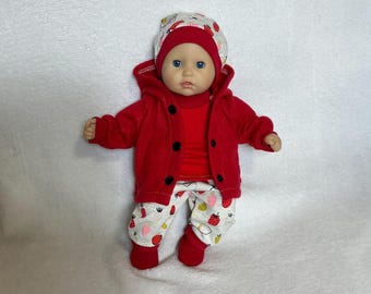May include: A baby doll with blue eyes wearing a red hooded jacket with black buttons, a red shirt, and white pants featuring a pattern of small red and pink strawberries. The doll also wears a matching white hat with a red brim and strawberry print.