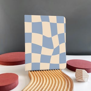 May include: A blue and cream checkered notebook with a wavy pattern, sitting on a wooden stand with a wavy design. The notebook is on a white surface with two red circles and a wooden geometric shape.