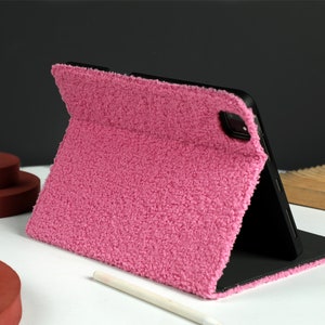 May include: A pink fuzzy tablet case with a black tablet inside. The case is folded to prop the tablet up for viewing.