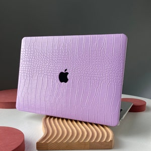 Puede incluir: Una funda para portátil de color lavanda con textura de cocodrilo en relieve y un logotipo de Apple. La funda está sobre un soporte de madera.