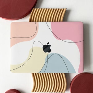 Puede incluir: Un portátil blanco con un diseño abstracto de colores en la tapa. El portátil está sobre un soporte de madera. Hay tres círculos rojos en el fondo.