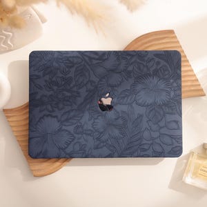 Puede incluir: Un portátil azul marino con un diseño floral. El portátil está colocado sobre una bandeja de madera. El logotipo de Apple es visible. También hay un frasco de perfume y objetos decorativos.