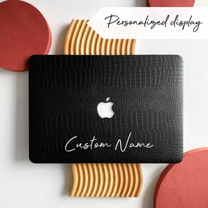 Puede incluir: Piel negra para portátil con un patrón de cocodrilo y un logotipo de Apple blanco. La piel tiene el texto "Custom Name" en letras blancas.