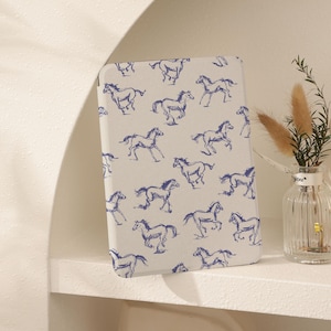 Può includere: Una copertina di taccuino rettangolare bianco sporco con un motivo ripetuto di illustrazioni di cavalli blu. I cavalli sono raffigurati in varie pose di corsa. Il taccuino è esposto su una mensola bianca, accanto a un piccolo vaso di vetro con piante decorative.