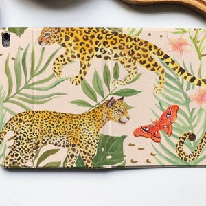 Puede incluir: Una funda para iPad rosa y beige con un estampado tropical con dos leopardos, hojas verdes y mariposas naranjas.