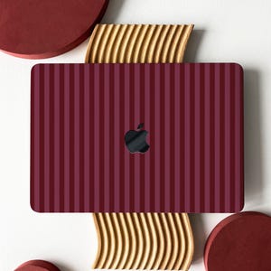 May include: A burgundy laptop skin with vertical stripes in varying shades of burgundy. The Apple logo is visible. The laptop is placed on a decorative wooden and maroon background.
