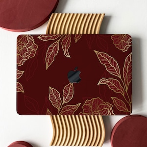 May include: A laptop computer with a burgundy red case featuring a gold floral design. The laptop is sitting on a wooden surface.