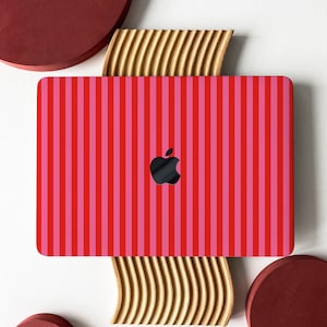 Pode incluir: Um laptop com um design listrado vermelho e rosa. O laptop apresenta um logotipo da Apple preto no centro. Ele está colocado em um suporte de madeira ondulado dourado, com objetos circulares vermelhos ao fundo.