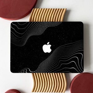 Puede incluir: Una funda negra para portátil con el logotipo blanco de Apple y un diseño de líneas onduladas blancas. La funda tiene un fondo moteado y se muestra sobre una superficie blanca con elementos decorativos de madera y granate.