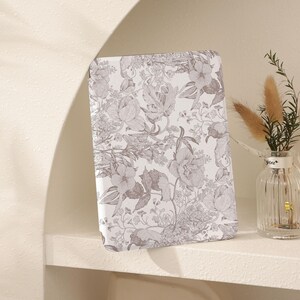 May include: A white book cover with a floral pattern in shades of gray. The cover is on a white shelf with a small vase of dried flowers.