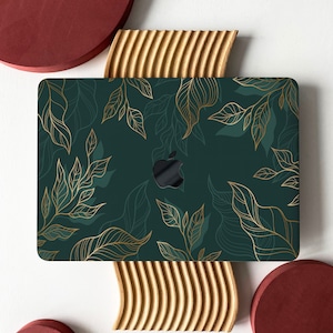 Könnte beinhalten: Eine grüne Laptop-Haut mit einem goldenen Blatt-Muster. Die Haut liegt auf einer Holzoberfläche mit einem roten Kreis auf jeder Seite.