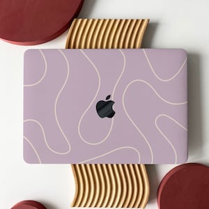 Puede incluir: Un portátil de color lila claro con un diseño abstracto blanco y un logotipo de Apple. El portátil está sobre un soporte de madera con un patrón ondulado. Hay dos círculos rojos a cada lado del portátil.