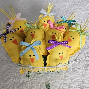 May include: A metal basket filled with handmade yellow chick ornaments. Each chick has a stitched orange beak, black eyes, and a colorful ribbon bow. The chicks are decorated with yellow fringe and are set against a white quilted background.