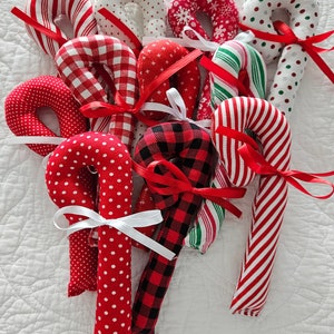 May include: A collection of handmade fabric candy canes in various patterns, including red and white stripes, polka dots, and plaid. Each candy cane is tied with a red or white ribbon, creating a festive holiday display.