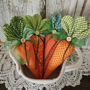 May include: A white teacup-shaped basket filled with fabric carrots. The carrots are orange with green fabric tops, wooden button accents, and green ribbon. The basket sits on a white lace doily, with a white wood background.