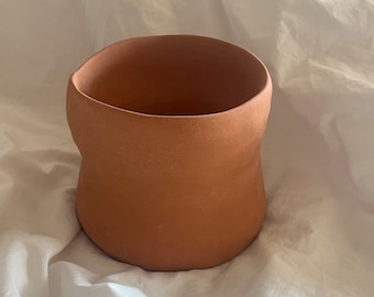 May include: A terracotta planter with a wide, rounded opening and a slightly constricted middle section. The planter has a rough, textured surface and a warm, earthy color.