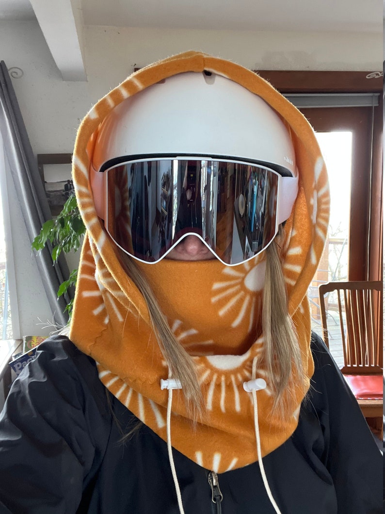 May include: A close-up of a person wearing a brown fleece neck gaiter with a white sun pattern. The gaiter has a drawstring closure and is worn over a white ski helmet with a pair of black ski goggles.