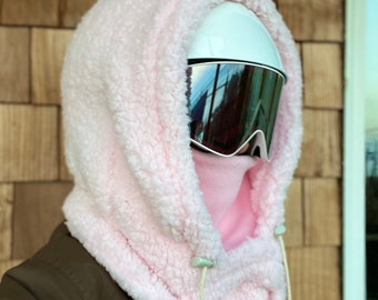 Puede incluir: Un pasamontañas de cuello de forro polar rosa con un cordón de ajuste blanco. El pasamontañas se lleva sobre un casco blanco y gafas de esquí negras.