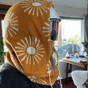 May include: A mustard yellow fleece neck gaiter with a white sunburst pattern. The gaiter has a white drawstring and is pulled up over the head.