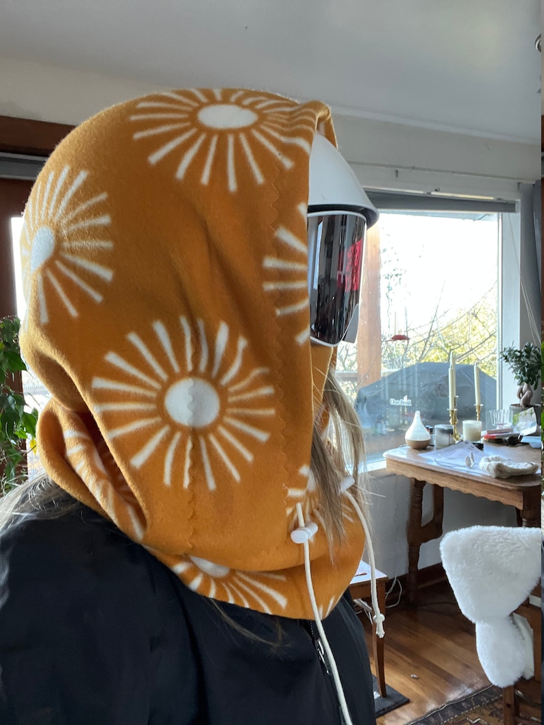 May include: A mustard yellow fleece neck gaiter with a white sunburst pattern. The gaiter has a white drawstring and is pulled up over the head.
