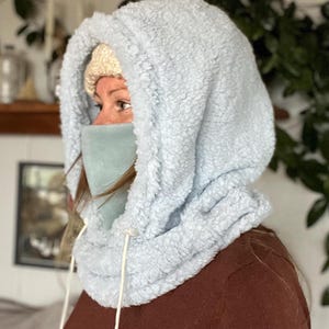 May include: A light blue hooded neck warmer with a white drawstring. The neck warmer is made of a soft, fluffy material and is worn by a person wearing a brown shirt.