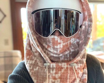 May include: A white ski helmet with large mirrored goggles worn with a fleece balaclava hood. The hood features a repeating geometric pattern in shades of terracotta, brown, and white, with two white drawstrings at the front neck area.