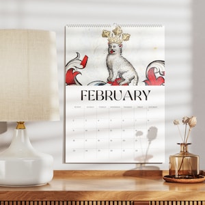 May include: A wall calendar for February featuring a regal cat illustration wearing a crown. The calendar is white with black text and hangs above a wooden surface. A white lamp and vase are also visible.