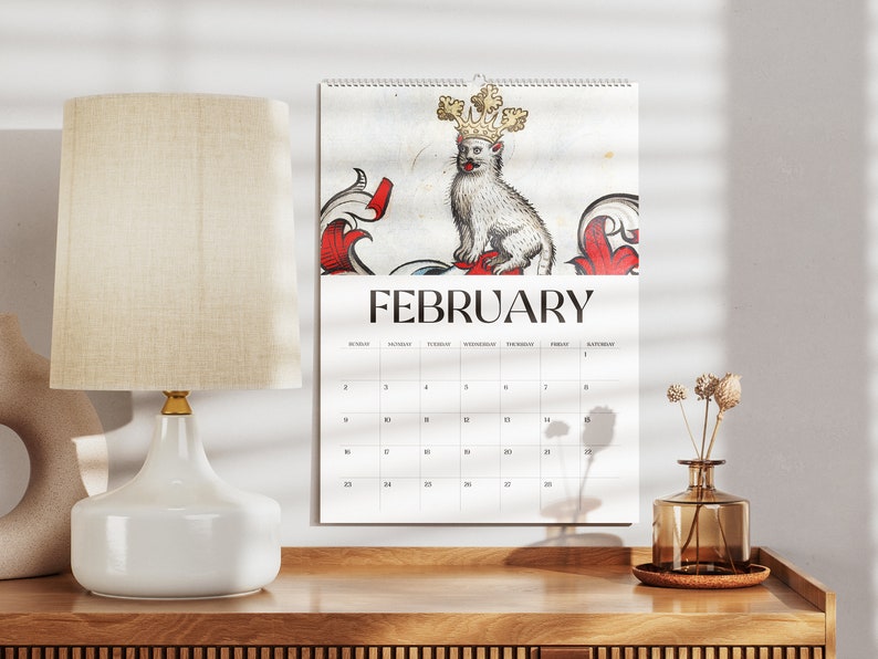May include: A wall calendar for February featuring a regal cat illustration wearing a crown. The calendar is white with black text and hangs above a wooden surface. A white lamp and vase are also visible.