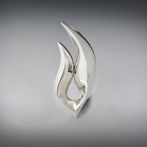 May include: A silver abstract sculpture with a flame-like design. The sculpture is made of smooth, polished metal and has a modern, minimalist aesthetic.