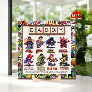 May include: A wooden sign with a comic book theme, featuring the word "DADDY" spelled out with Scrabble tiles. Below are cartoon superhero characters with descriptive phrases. The sign includes the text "YOU ARE OUR SUPERHERO! LOVE, LUCY AND NOAH."