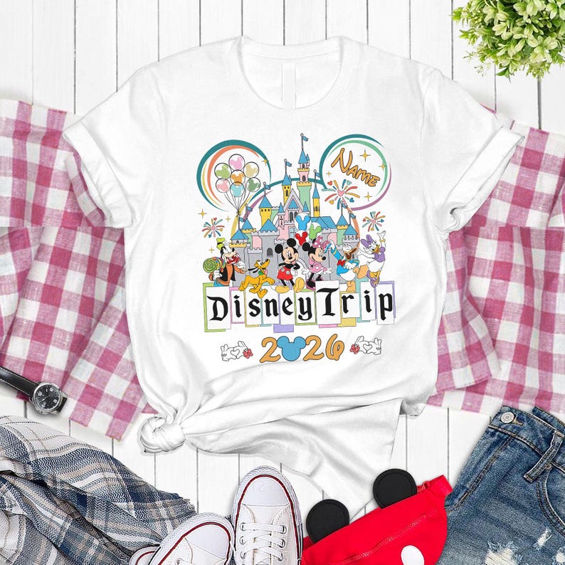 A white t-shirt with a Disney-themed graphic design, featuring Mickey Mouse, Minnie Mouse, Donald Duck, and other Disney characters, along with the text "Disney Trip" and "2026". The t-shirt is displayed on a white background, surrounded by various clothing items and accessories.