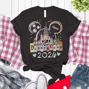A black t-shirt with a Disney Land-themed design, featuring Mickey Mouse and Minnie Mouse characters, along with the text "Disneyland 2020". The t-shirt is displayed on a wooden surface, surrounded by various clothing items such as jeans and sneakers.