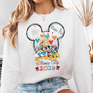A white sweatshirt with a colorful graphic design featuring Disney characters and the text "Disney Trip 2020".