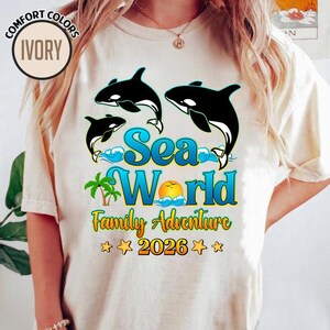 May include: Ivory-colored t-shirt featuring a graphic of three black and white orcas leaping above blue waves. The text "Sea World Family Adventure 2026" is displayed in blue and yellow, with palm trees and a sun graphic.