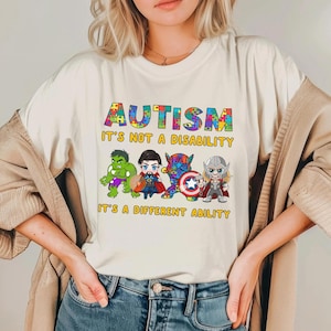 May include: Cream-colored t-shirt with the word "AUTISM" in colorful puzzle piece letters. Below the word is the phrase "IT'S NOT A DISABILITY IT'S A DIFFERENT ABILITY". The shirt features cartoon illustrations of superheroes.