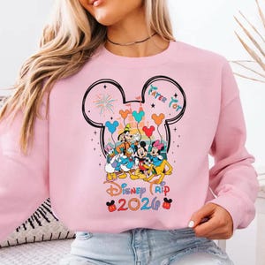 A woman wearing a pink sweatshirt with a Disney-themed graphic design featuring Mickey Mouse and other Disney characters.