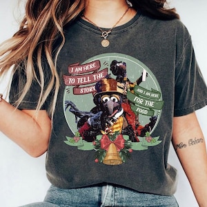 May include: Dark gray t-shirt featuring a graphic of Gonzo and other Muppet characters. The design includes text that reads "I AM HERE TO TELL THE STORY" and "AND I AM HERE FOR THE FOOD". A festive bell and ribbon are also included in the design.