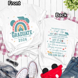 a white t-shirt with a colorful graphic and text, laid out on a wooden surface with a plaid blanket and a pair of sneakers.