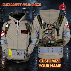 May include: A beige zip-up hoodie featuring a printed Ghostbusters uniform design. The front displays a name tag reading STANTZ and a belt graphic. The back shows a detailed proton pack illustration with tubes, wires, and a ghost logo on the sleeve.