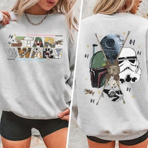 May include: A light gray sweatshirt featuring the text "STAR WARS" in a colorful, stylized font on the front. The back of the sweatshirt displays iconic Star Wars characters and elements, including the Death Star and a Stormtrooper helmet.