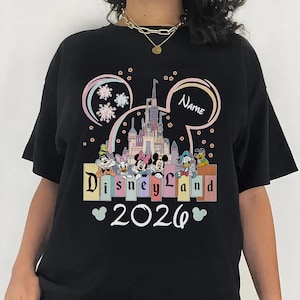 A black t-shirt with a Disney Land theme, featuring a castle, Minnie Mouse, Mickey Mouse, Donald Duck, and other Disney characters. The text "Disneyland" is displayed prominently, and the year "2026" is shown below.