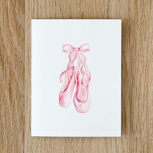 Peut inclure: Une carte de vœux blanche présentant une illustration à l'aquarelle centrée d'une paire de chaussons de danse roses attachés ensemble par un nœud en ruban rose assorti. L'illustration utilise des tons rouges et roses doux pour les chaussons.