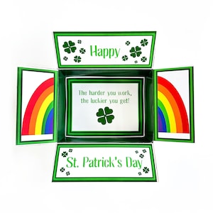 Può includere: Un biglietto verde e bianco con un disegno di trifoglio e il testo "Happy St. Patrick's Day". Il biglietto è piegato a forma di scatola e ha un disegno arcobaleno all'interno.