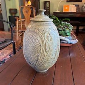 May include: A large, oval-shaped ceramic jar with a lid, featuring a textured, carved design. The jar is a muted, earthy tone, likely stoneware. It sits on a dark wooden table, showcasing its size and craftsmanship. The jar is approximately 18 inches tall.