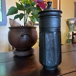 May include: A dark gray, cylindrical decorative jar with a lid, featuring intricate embossed patterns. The jar is placed on a dark wooden table, with a potted plant in the background. The jar's design includes vertical and geometric elements.