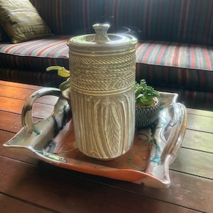 May include: A decorative ceramic jar with a lid, featuring a textured design, sits on a colorful tray with handles. A small potted plant is also on the tray. The tray is placed on a wooden table.