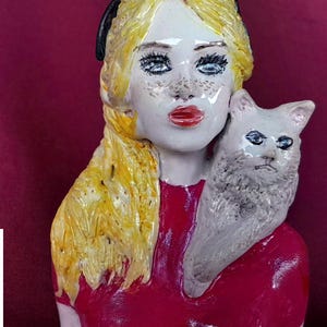 Puede incluir: Escultura de cerámica de una mujer con cabello rubio y vestido rojo, sosteniendo un gato blanco. La mujer tiene labios rojos, ojos azules y pecas. El gato tiene una expresión severa. La escultura está sobre un fondo burdeos.