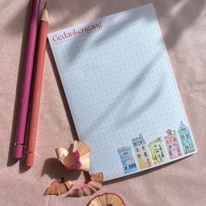 Può includere: Un blocco note bianco con un motivo a puntini blu e il testo "Gedanken gang" in alto. Il blocco note ha una fila di case colorate disegnate in basso. Due matite rosa e trucioli di matita sono su una superficie rosa chiaro.
